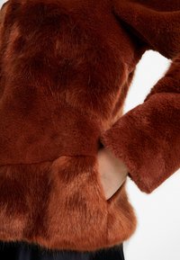 Main dans la poche d'une veste brune en fourrure avec des manches longues, sur un fond clair uni.