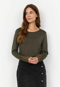 Olivgrön långärmad topp med rund halsringning och intrikata spetsdetaljer på ärmarna, tillsammans med en svart denimkjol med knappar.