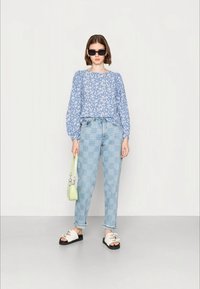 Blusa azul com padrão floral, jeans xadrez azul claro, sandálias planas pretas com tiras brancas e uma pequena mala verde.