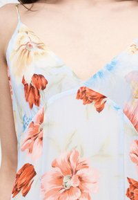 Γυναίκα που φοράει ανοιχτό μπλε φόρεμα με λεπτούς ιμάντες και σχέδια από ροδακινί, κόκκινα και κίτρινα λουλούδια.