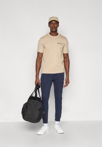 Μπεζ μπλουζάκι με λογότυπο, σκούρο μπλε φόρμα, λευκά αθλητικά παπούτσια και μια μαύρη τσάντα ντάφελ. Το μοντέλο φοράει καπέλο ανοιχτού χρώματος.