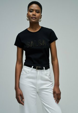 Femme portant un t-shirt noir à manches courtes avec le texte "SALSA JEANS" et un pantalon blanc taille haute, debout devant un fond gris uni.