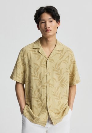 Giovane uomo con camicia giallo chiaro a maniche corte con motivo a foglia e pantaloni bianchi, in piedi con le mani in tasca.