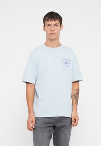 Jongeman met kort haar, gekleed in een lichtblauwe T-shirt met opdruk "Only & Sons Noodles" en een grijze spijkerbroek, staand tegen een witte achtergrond.