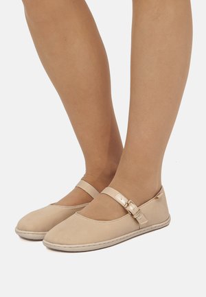 Piedi che indossano ballerine stile Mary Jane beige con una singola fascia e fibbia su uno sfondo bianco.