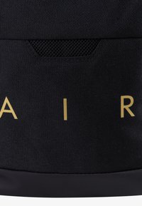 Bolsa de tela negra con letras doradas "AIR." Presenta una superficie texturizada, un bolsillo de malla y una base suave. Diseño simple y funcional.