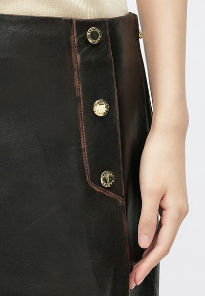 Brūna ādas svārki ar zelta pogām sānos, kontrastējoša dūriena un gluda tekstūra. Tuvplāns uz svārkiem un roku novietojumu.