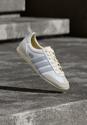 Weißer und hellblauer Sneaker mit Perforationen, beigen Schnürsenkeln und strukturiertem Gummisohlen. Verfügt über drei Streifen und goldene Markenkennzeichnung.