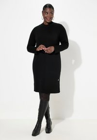 Robe pull noire en tricot avec des manches longues, texture côtelée, col rond et une accent doré à l'ourlet, associée à des bottes noires jusqu'aux genoux.