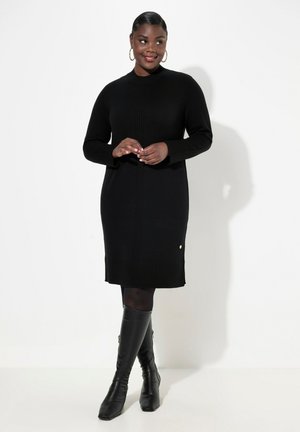 Schwarzes Strickkleid mit langen Ärmeln, geripptem Textil, rundem Halsauschnitt und einem goldenen Akzent am Saum, kombiniert mit schwarzen kniehohen Stiefeln.