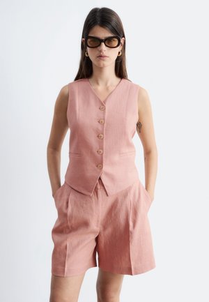 Frau trägt eine ärmellose rosa Knopfwest und passende Shorts, mit Sonnenbrille und Händen in den Taschen.