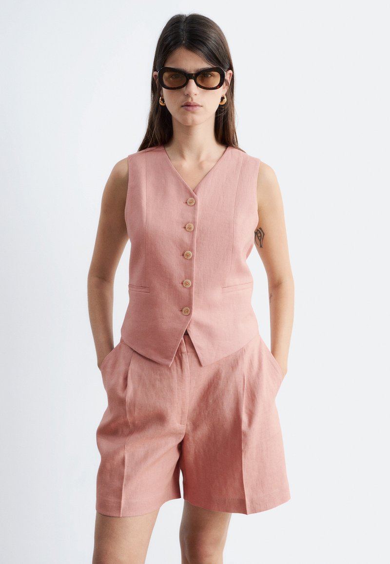 Mujer con chaleco rosa sin mangas con botones y pantalones cortos a juego, con gafas de sol y las manos en los bolsillos.
