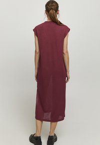 Robe sans manches bordeaux en tissu tricot léger et fluide ; longueur genou avec fentes sur les côtés. Texture douce et design simple et minimaliste.