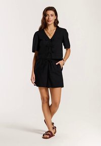 Zwart kort mouwshirt met een V-hals en een trekkoord aan de voorkant, gecombineerd met zwarte shorts met elastische taille. Draag gecombineerd met bruine sandalen.