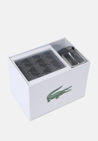Caixa de presente branca com tampa transparente, contendo um item com padrão preto e uma fivela prateada. Apresenta um logótipo de crocodilo verde na lateral.