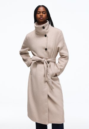 Femme aux longues tresses portant un manteau beige ceinturé avec un col montant et des boutons noirs, debout les mains dans les poches sur fond blanc.