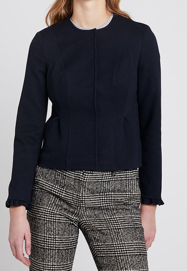 Námořnická modrá cropped bunda s kulatým výstřihem, texturovanou tkaninou a jemnými detaily švů; spárovaná s kostkovanými, taperovanými kalhotami s volánovými manžetami.