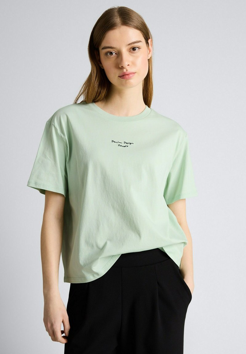 Jonge vrouw met steil bruin haar, gekleed in een lichtgroen T-shirt met kleine zwarte tekst en een zwarte broek, staand tegen een effen achtergrond.