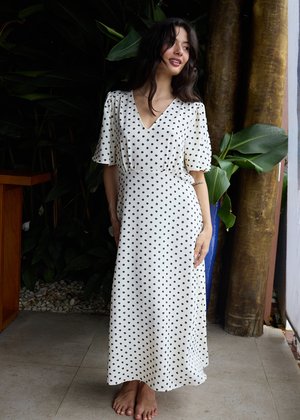 Kobieta w białej sukience midi w czarne grochy stoi boso w pomieszczeniu obok dużych zielonych roślin i drewnianych mebli, patrzy na bok i uśmiecha się.