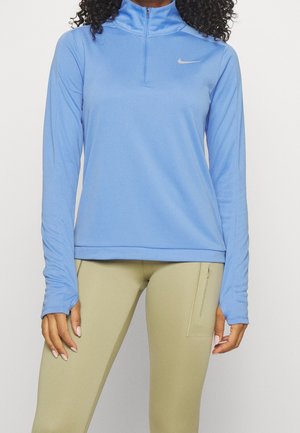 Kobieta ubrana w jasnoniebieską sportową bluzę z długim rękawem i półzamkiem oraz beżowe legginsy z suwakiem w bocznej kieszeni, stojąca na jednolitym tle.