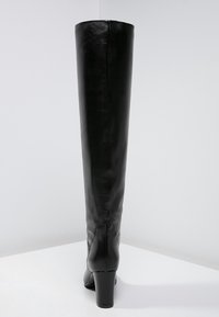 Bottes noires en cuir montantes avec une texture lisse, un talon bloc haut et des détails de couture le long de l'arrière.