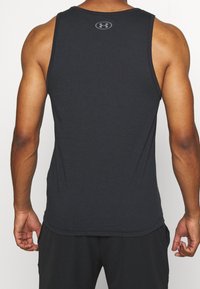 Vahva henkilö pukeutunut mustaan ilmanhihaa urheilupaitaan, jossa on Under Armour -logo yläselässä, seisoo yksinkertaista taustaa vasten.