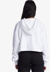 Sudadera corta blanca con capucha ajustable, puños acanalados y corte holgado, combinada con pantalones negros de cintura alta. Textura de tela suave.