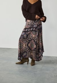 Jupe longue à motifs avec un fond sombre présentant un motif paisley en rose, bleu et or. Associée à des bottes à talons vert olive.