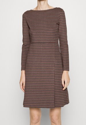 Kobieta w sukience za kolano z długim rękawem o drobnym wzorze w brązowo-czarną pepitkę, stojąca na jednolitym jasnym tle.