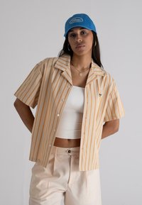 Short-sleeve shirt in cream with orange vertical stripes, paired with a white tank top and beige pants, complemented by a blue cap.