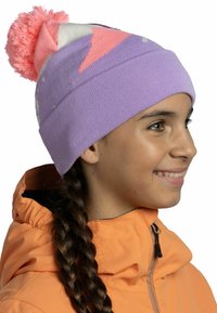 Purple knit beanie with a pink pom-pom on top, featuring geometric patterns in pink and white. Worn with an orange jacket.