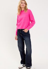 Fuchsienfarbener Strickpullover mit langen Ärmeln und Rundhalsausschnitt, kombiniert mit dunkelblauen Jeans mit weitem Bein und weißen Sneakers mit schwarzen Streifen.