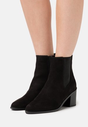Bottes en daim noir à cheville avec bout pointu, panneaux latéraux élastiques et talon carré solide. Finition texturée avec des accents de coutures nettes.