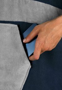 Graues und marineblaues strukturiertes Material mit einer großen Tasche; eine Hand legt ein blaues Smartphone von der Seite in die Tasche.