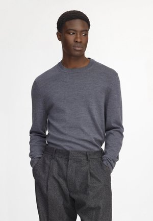 Ung mand iført en tætsiddende grå langærmet sweater og højtaljede mørkegrå plisserede bukser med hænderne i lommerne.