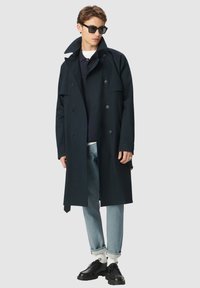Granatowy płaszcz trench z podwójnymi guzikami, dużym kołnierzem i kieszeniami; w połączeniu z granatowym polo, jasnoniebieskimi dżinsami i czarnymi butami.