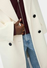 Witte wollenmix jas met een notched kraag, zwarte knopen en zijzakken, gedragen over een donkerrode zip-up trui en blauwe spijkerbroek.