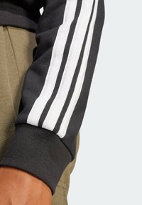 Svart sweatshirt med vita och grå randiga detaljer på ärmarna. Har ribbade muddarna och en mjuk textur, visad mot olivgröna byxor.