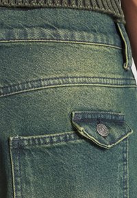 La tela de mezclilla verde cuenta con un bolsillo con cierre de botón de metal. La textura muestra un leve desgaste y detalles de costura a lo largo de las costuras.