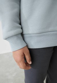 Lyseblå ribbet sweatshirt med tætsiddende manchetter og talje, vist med en hånd hvilende på mørkegrå joggingbukser i en neutral indstilling.