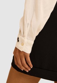 Blusa bianca con texture e maniche arricciate, caratterizzata da un fiore ricamato nero e un polsino con bottoni dorati, abbinata a un indumento nero.