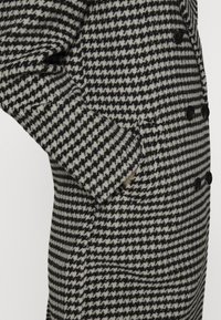 Abrigo de doble botonadura con un patrón de espiga en blanco y negro, fabricado en una tela texturizada con botones negros y bolsillos laterales.