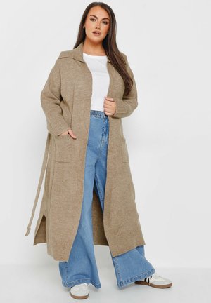 Larga chaqueta beige con cuello, aberturas laterales y dos bolsillos frontales, combinada con jeans de pierna ancha de tiro alto en azul y zapatillas blancas.
