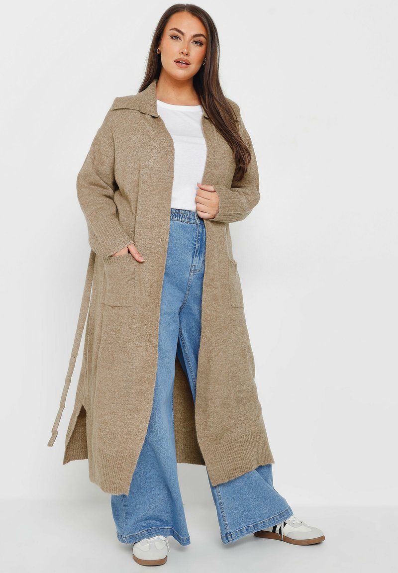 Lång beige kappa med krage, sidofickor och två framfickor, tillsammans med högmidjade blå vida jeans och vita sneakers.