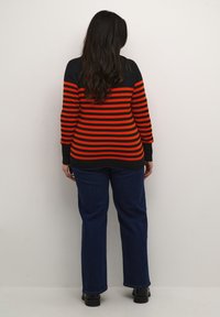 Pull rayé noir et orange à manches longues et col rond, associé à un jean large bleu foncé et des chaussures noires.