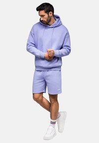 Haut en coton lilas avec poche kangourou, associé à un short assorti. Le modèle porte des baskets blanches et des chaussettes à rayures à la cheville.