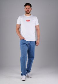 Weißes Baumwoll-T-Shirt mit einem roten rechteckigen Logo auf der Brust, kombiniert mit hellblauen Jeans und weißen Sneakern. Einfaches, lässiges Outfit.