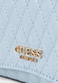 Borsa imbottita azzurro chiaro con un motivo testurizzato, accessori in tonalità oro e logo "GUESS EST 1981" in evidenza.