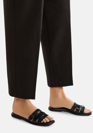 Černé pantofle s více pásky, plochou podrážkou a obdélníkovým tvarem; nošené s širokými černými zkrácenými kalhotami. Hladká kůže.