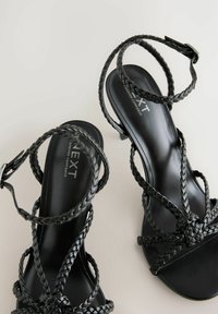 Schwarze Sandalen mit Riemchen, die geflochtene Lederstraps, ein glattes Fußbett und einen moderaten Absatz aufweisen. Das Design beinhaltet eine Schnallenverschluss zur Anpassungsfähigkeit.